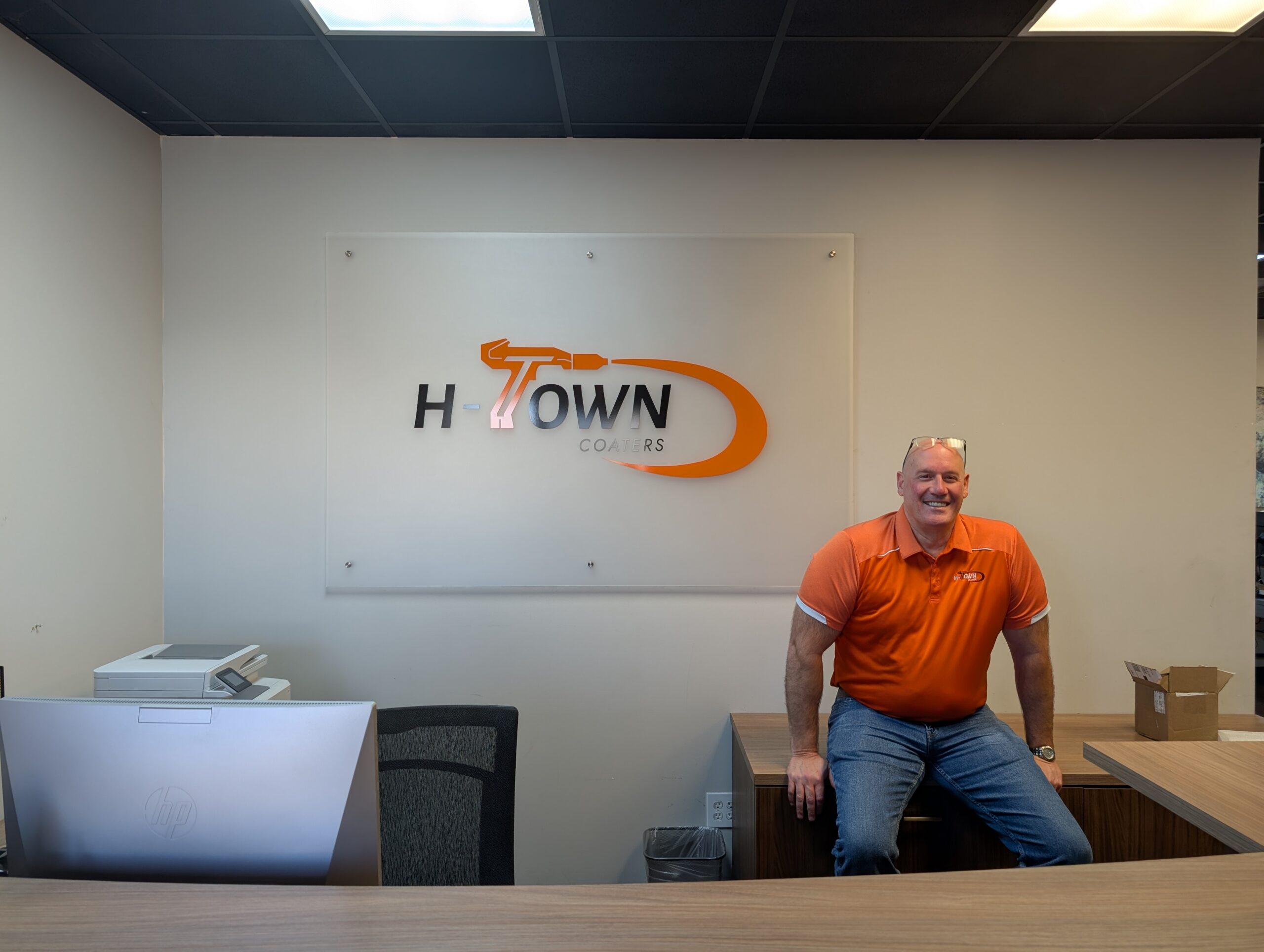 H-Town Coaters owner Jarrod Hammer seated in the front office beside the company logo in Houston, Texas — representing trusted local powder coating expertise and personalized service.